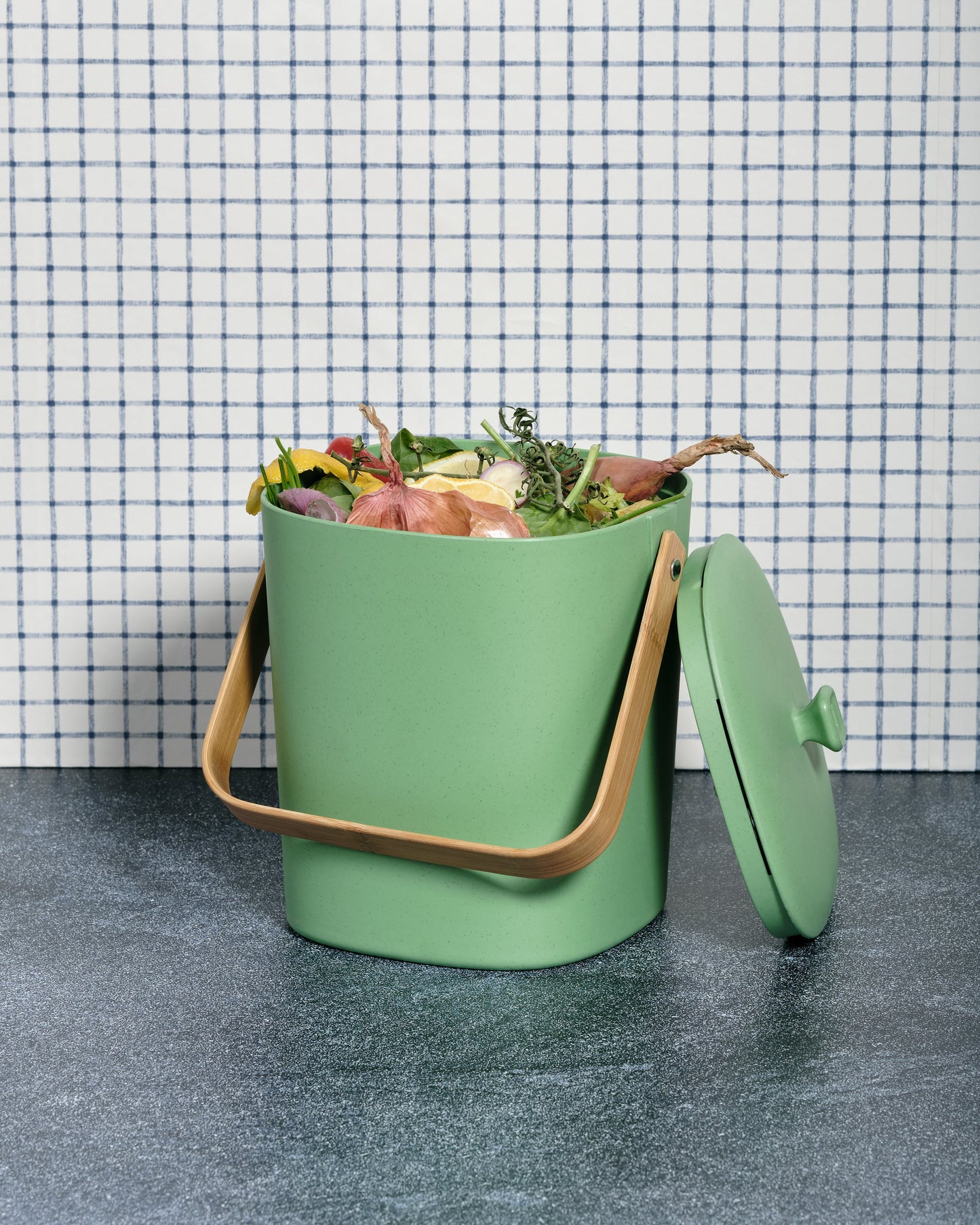 Avocado-colored Astrik Compoast Bin made of 100% biodegradable plant-based plastic with a bamboo handle on a gray counter with a tiled wall.