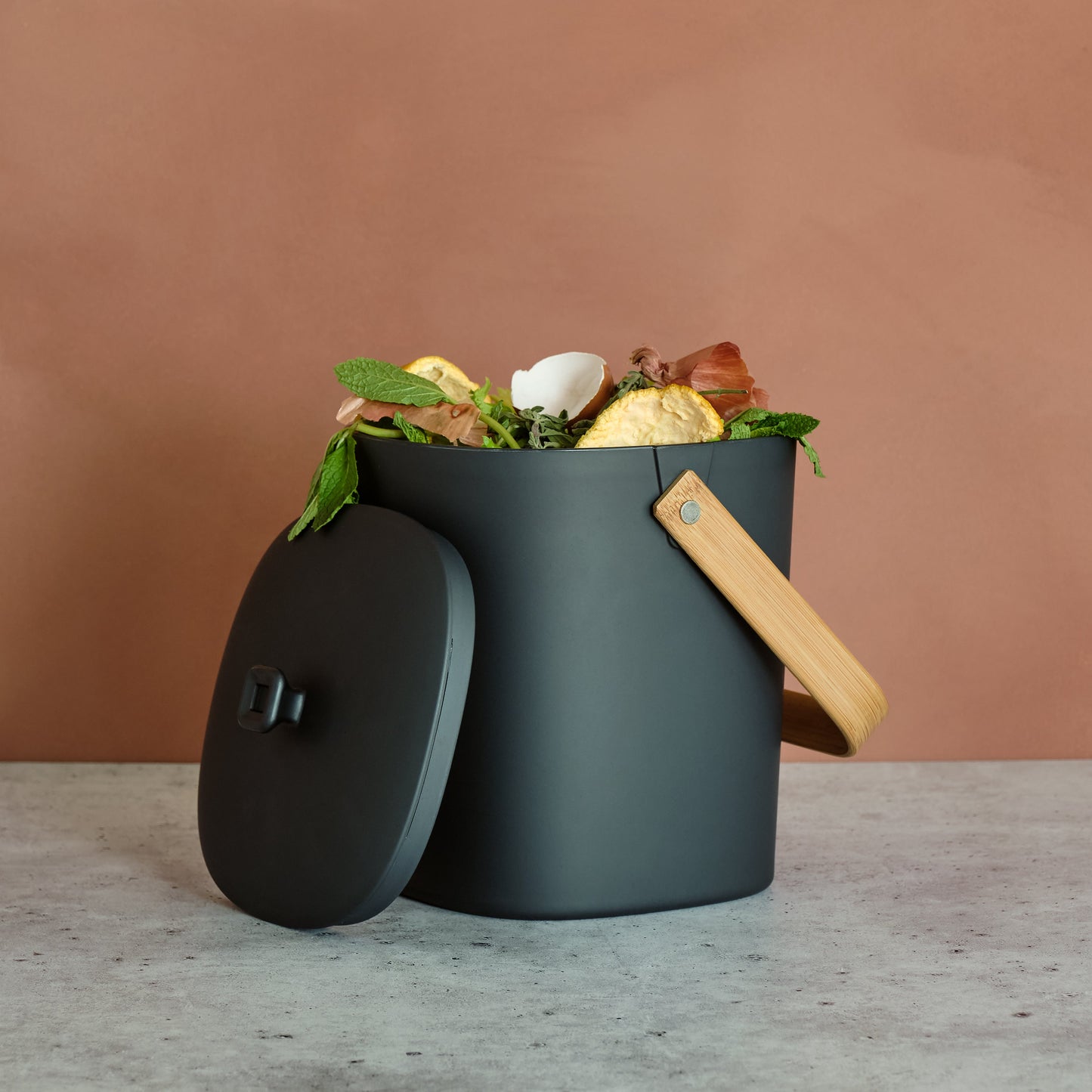 Graphite-colored Astrik Compoast Bin made of 100% biodegradable plant-based plastic with a bamboo handle on a gray counter with a salmon-colored background.