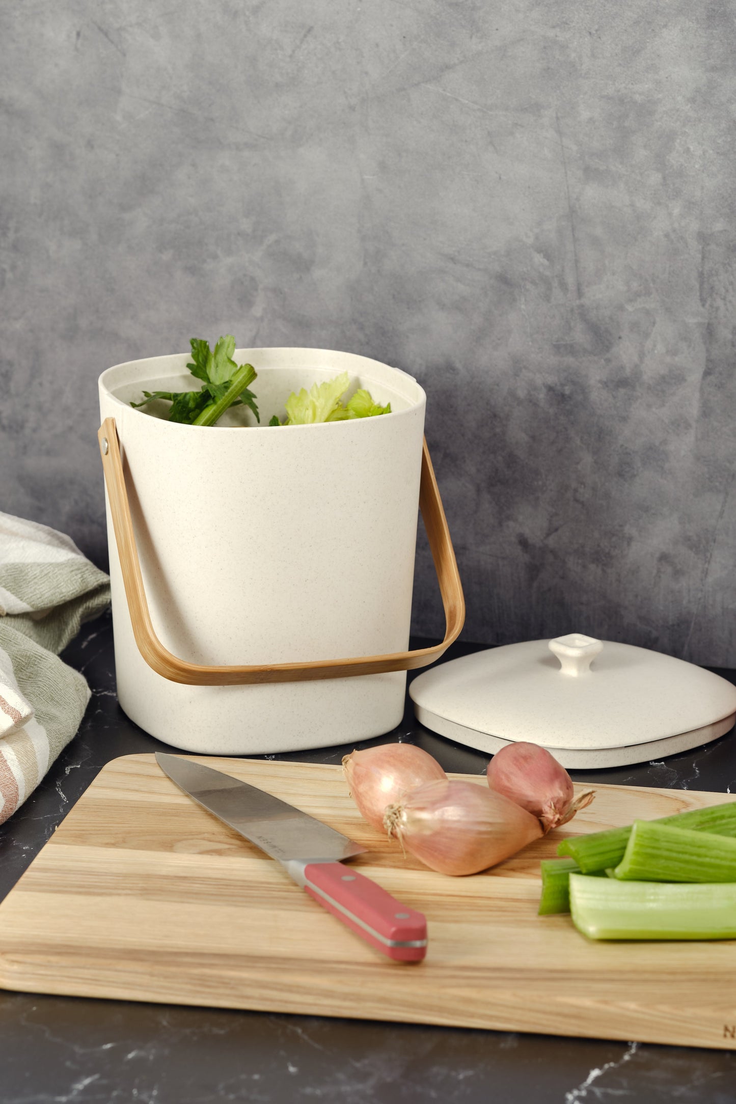 Natural-colored Astrik Compoast Bin made of 100% biodegradable plant-based plastic with a bamboo handle on a gray counter alongside a cutting board with vegetables.