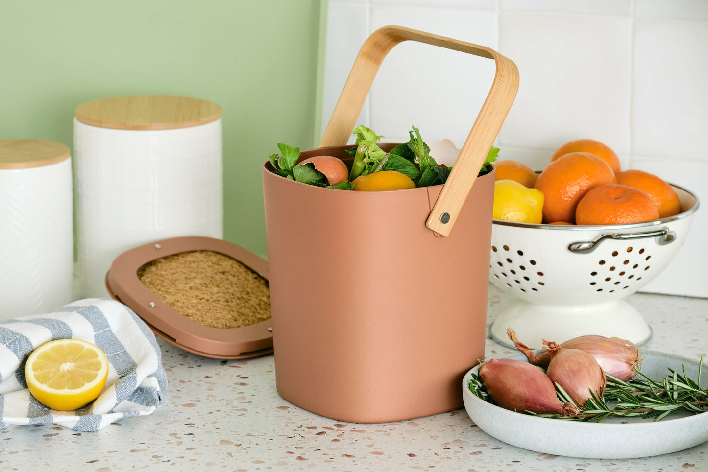 Terracotta-colored Astrik Compoast Bin made of 100% biodegradable plant-based plastic with a bamboo handle on a kitchen counter with fruits, cannisters herbs and onions.