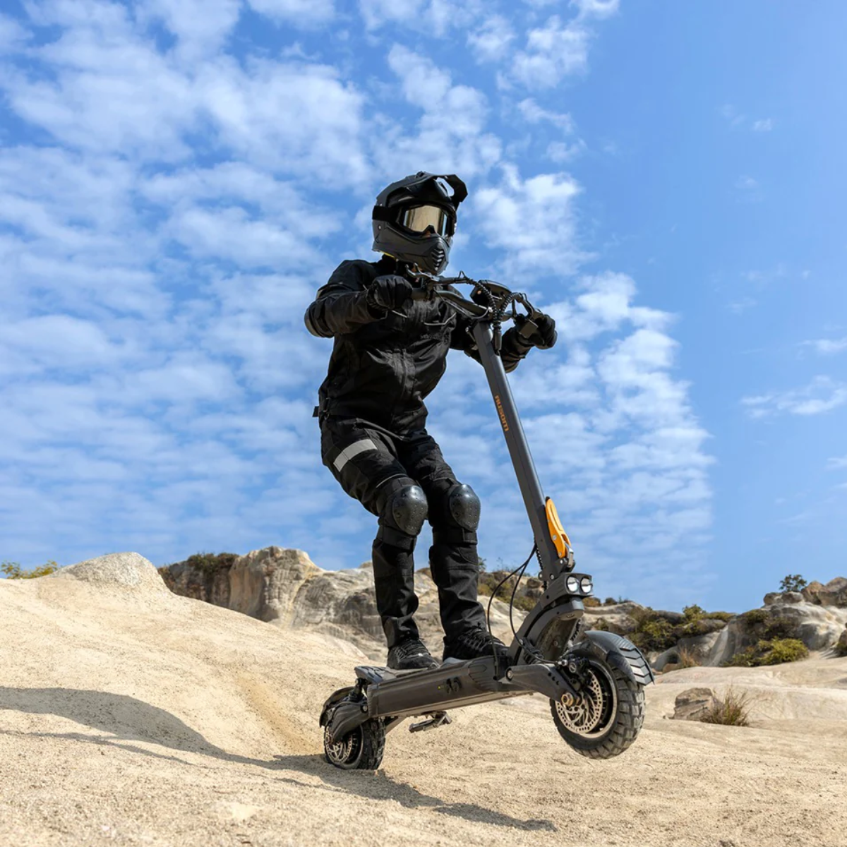 Person in full protective gear riding an AUSUM F1 Max electric scooter on a rocky landscape with a blue sky.
