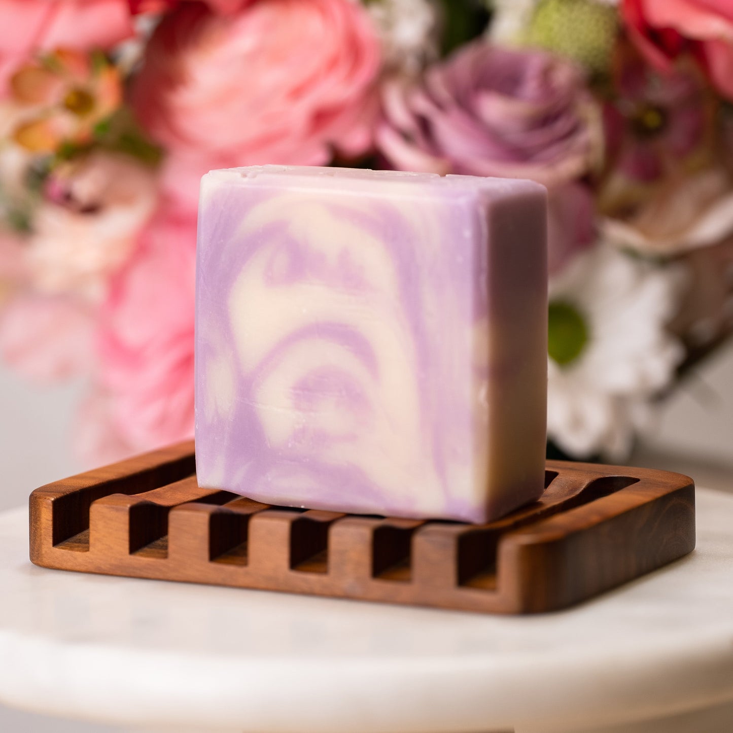 Swirled purple and white lavender soap bar on a wooden soap dish with flowers in the background