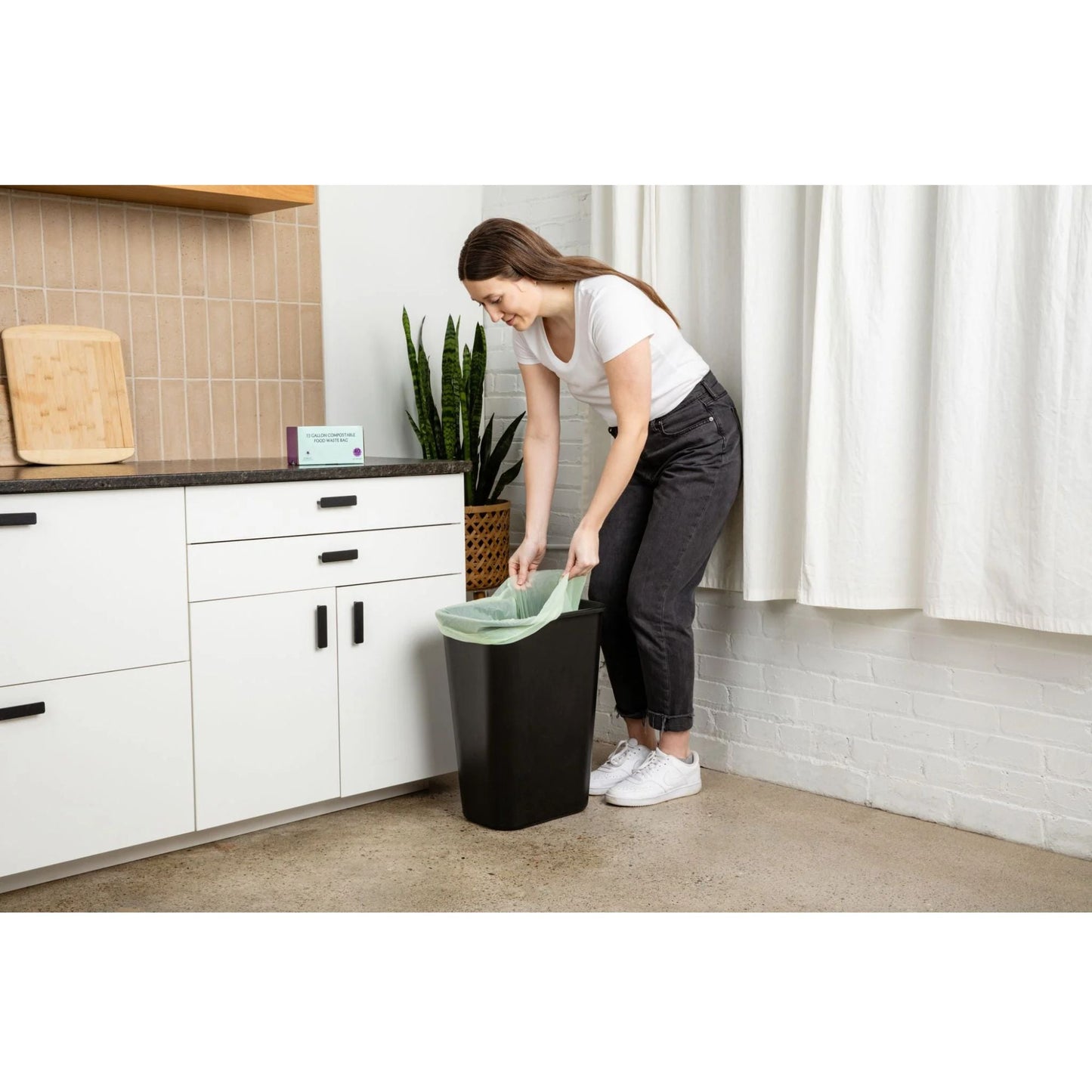 Woman in a kitchen placing a green 13 gallon compostable garbage bag into a black trash can.