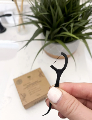 Black biodegradable dental floss picks held by a hand with a plant and kraft packaging box in the background