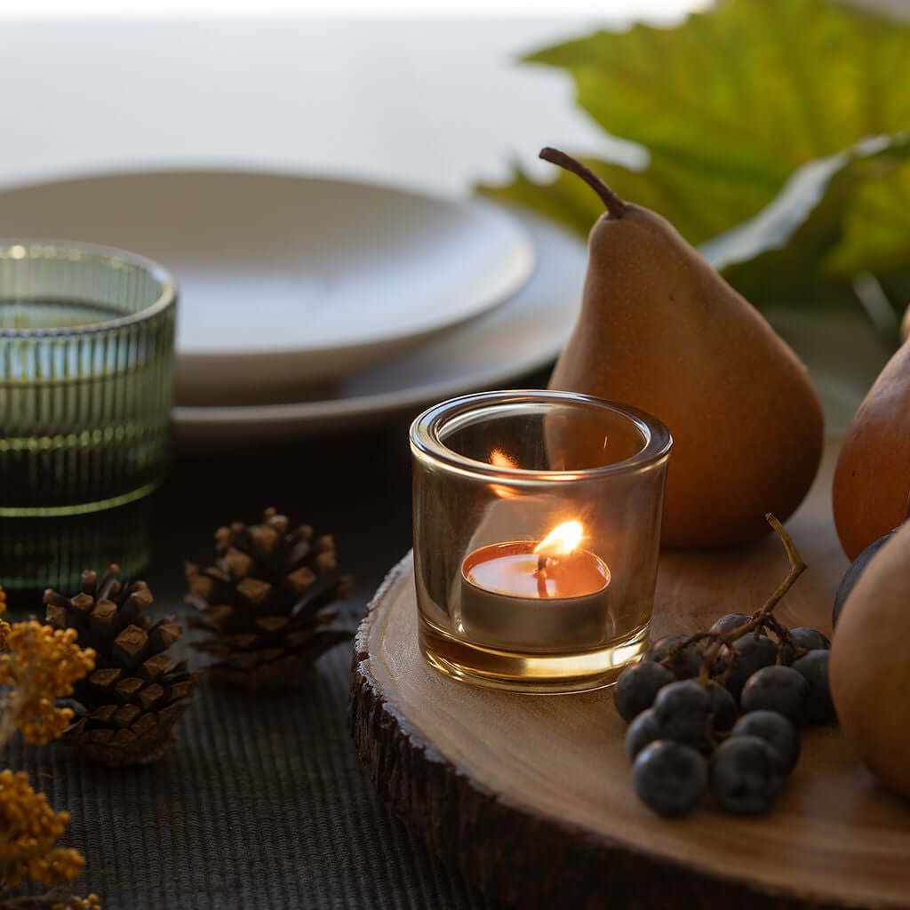 Bluecorn Pure Beeswax Tea Light Candle in a small glass holder on a wooden board with fruits and leaves.
