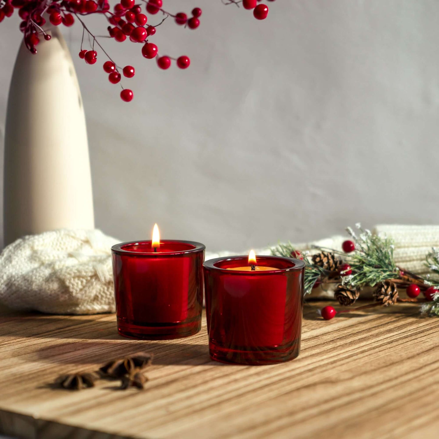 Two red candles on a wooden surface with a vase and decorative elements in the background.