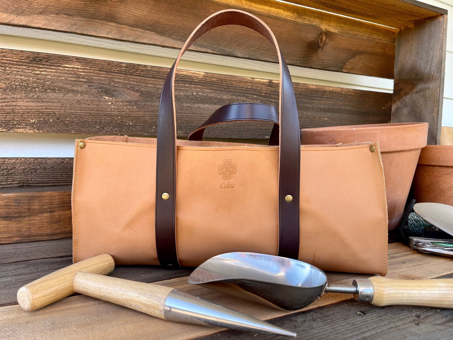 Brown leather tote bag with handles on a wooden surface with master garden gift set tools.