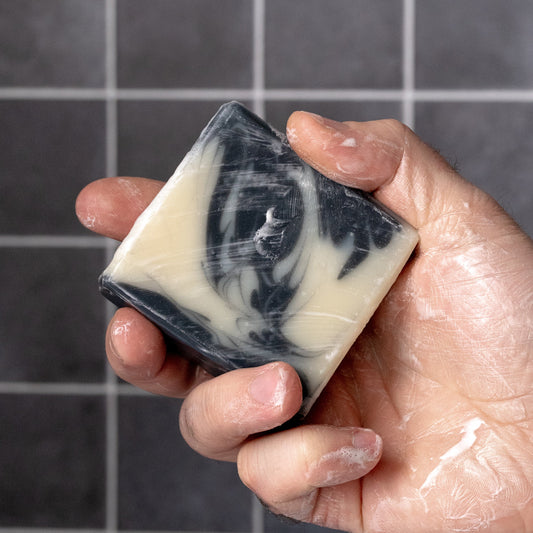 Hand holding a bar of detoxifying charcoal soap with black and white swirls against a tiled wall background