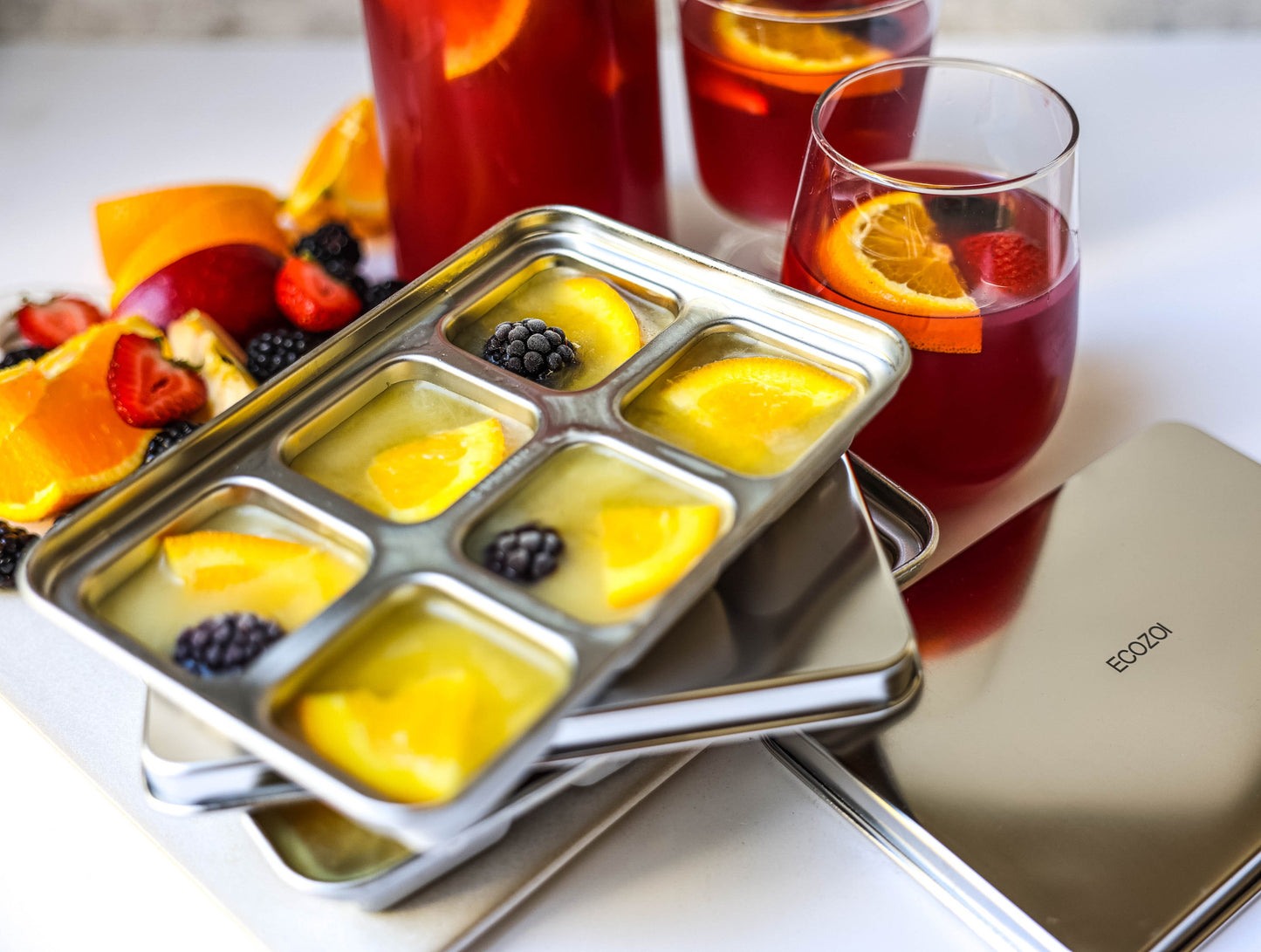 Eco-friendly stainless steel ice cube tray with fruit pieces next to glasses of red liquid on a white surface.