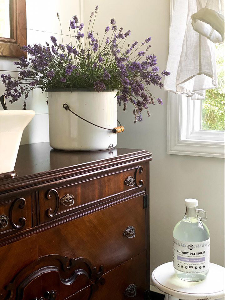 White bucket with lavender flowers on a wooden dresser next to a bottle of lavender detergent.