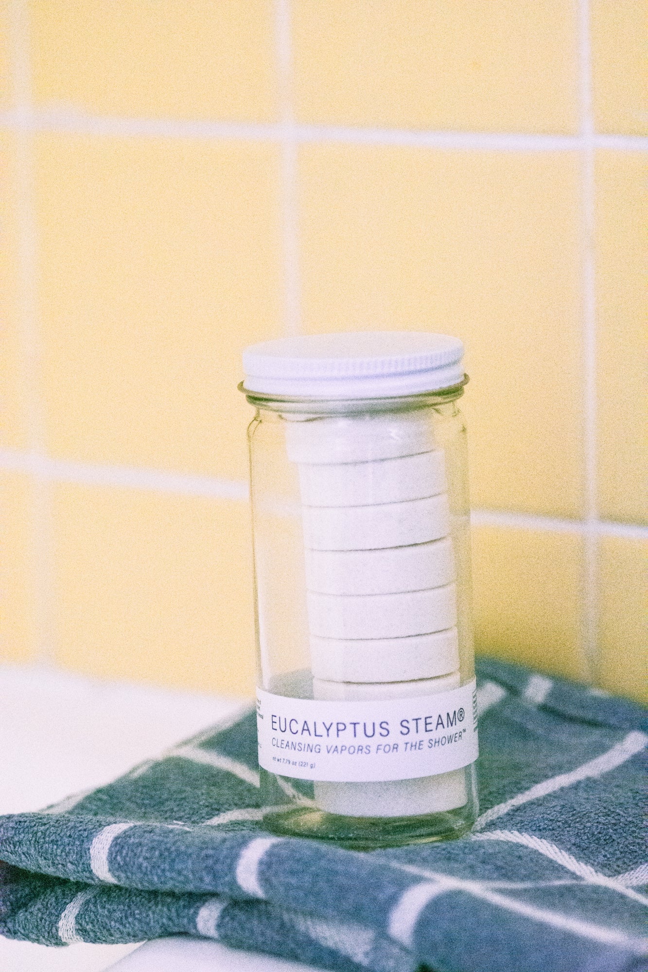 Jar of eucalyptus steam shower vapors by No Tox Life on a towel with a yellow tiled wall background