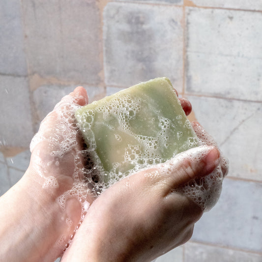 Hand holding a bar of peppermint tea tree soap with foam against a tiled bathroom wall.
