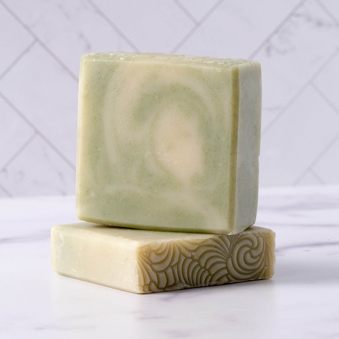 Two peppermint tea tree soap bars with a swirled pattern on a white surface with a light gray geometric background