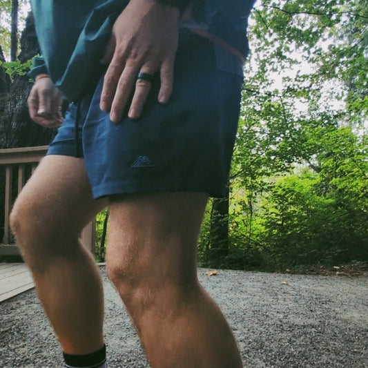 Person wearing Athletic 5 Inch Recycled Shorts in Cascade Blue by Rainier Watch, shorts showing a logo in a forest setting
