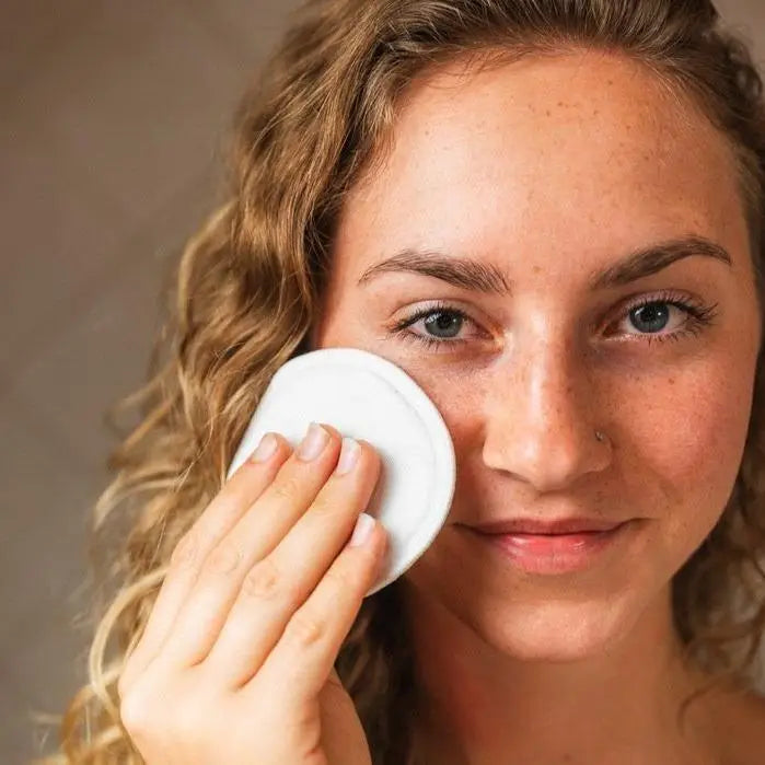 Woman applying a white bamboo facial round to her face against a neutral background