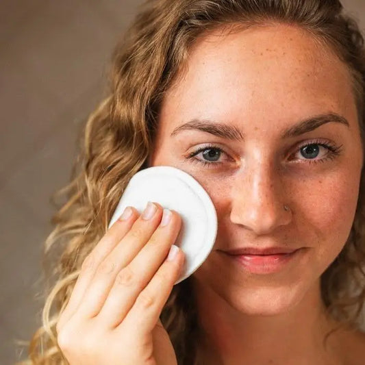 Woman applying a white bamboo facial round to her face against a neutral background
