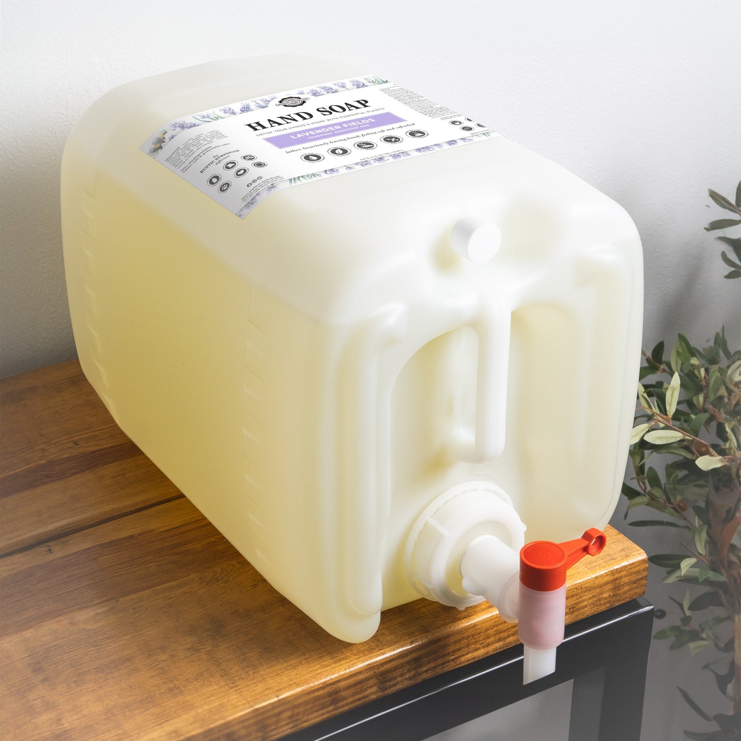 5-gallon plastic container of Rustic Strength Lavender Fields Liquid Hand Soap on a wooden surface with a plant in the background