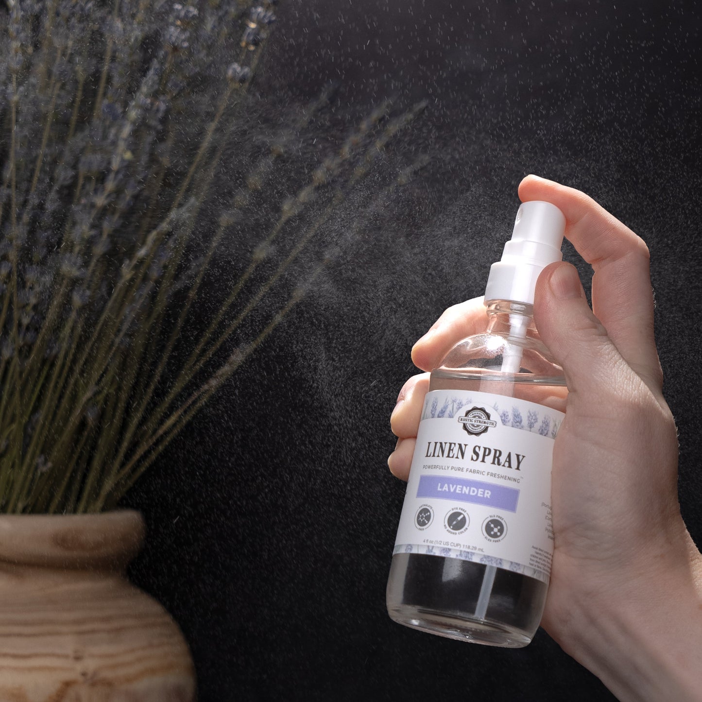 Hand holding a bottle of Rustic Strength Linen Spray, Lavender scented, with a dark background and lavender plant.