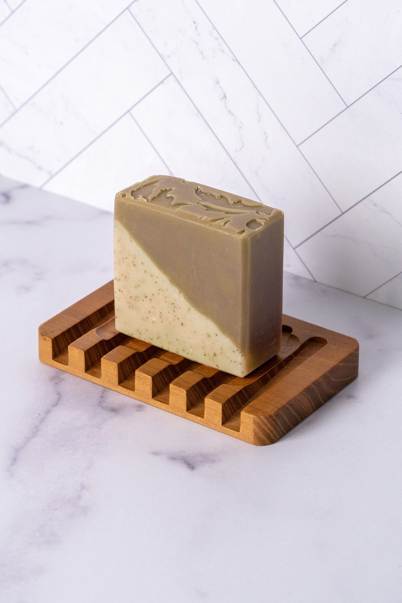 Bar of sandalwood patchouli soap on a wooden soap dish against a white tiled wall.