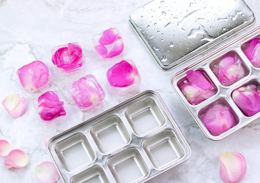 Stainless steel ice cube trays with pink rose petals in ice cubes on a marble surface
