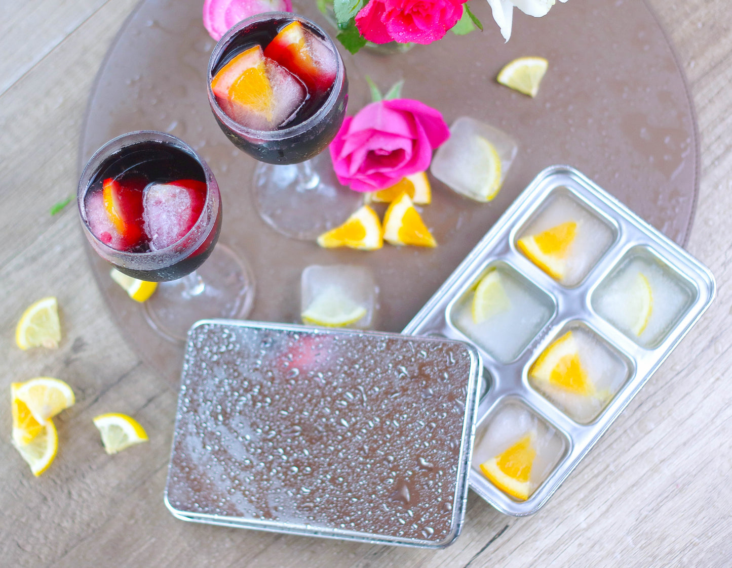Stainless steel ice cube tray with lid filled with lemon ice cubes and glasses of red wine with fruit, and flowers on a wooden surface.