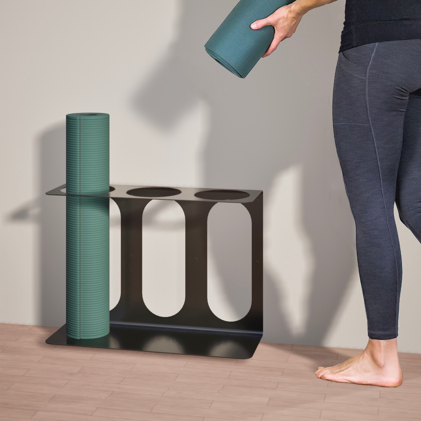 Person holding a yoga mat next to a black Yoke yoga mat wall bracket, suited to fit up to 3 yoga mats or rollers with rolled-up mat stowed in a slot, on a wooden floor.