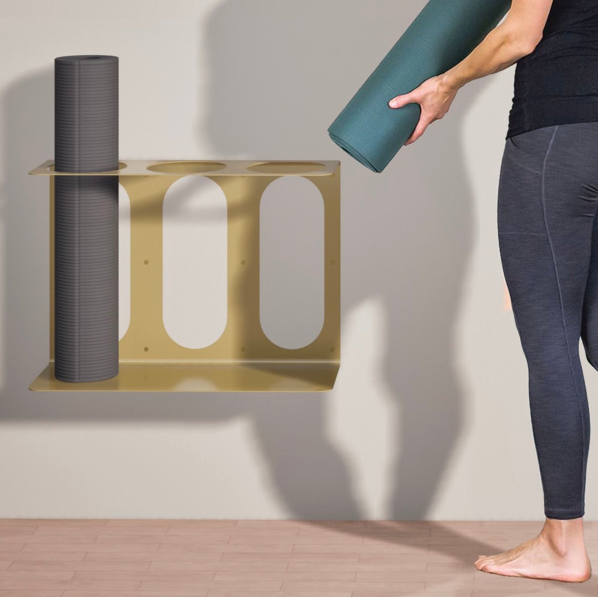 Person holding a yoga mat next to a gold Yoke Yoga Mat Wall Bracket, suited to fit 3 yoga mats or rollers on a neutral background