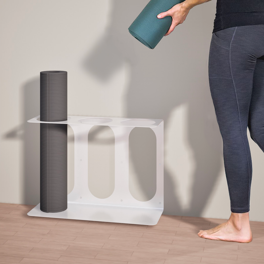 Person holding a yoga mat next to a white Yoke yoga mat wall bracket that fits 3 yoga mats or rollers on a wooden floor.