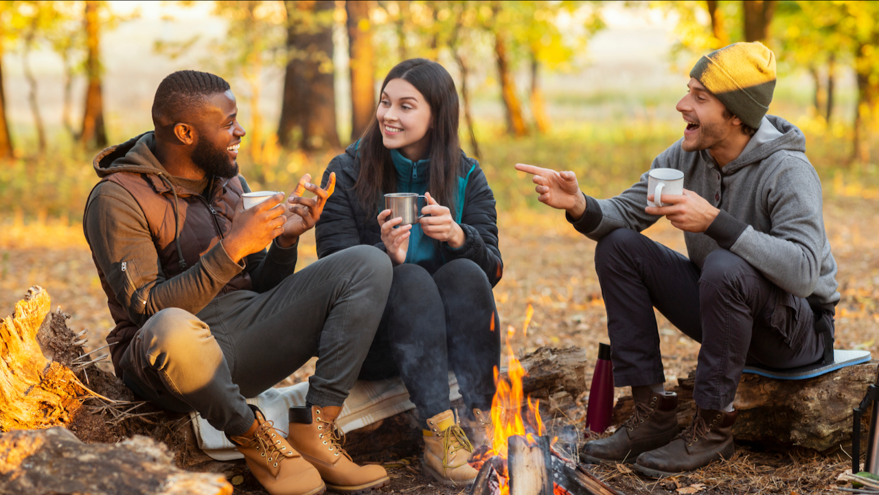 Friends laughing at an outdoor fire with a warm beverage