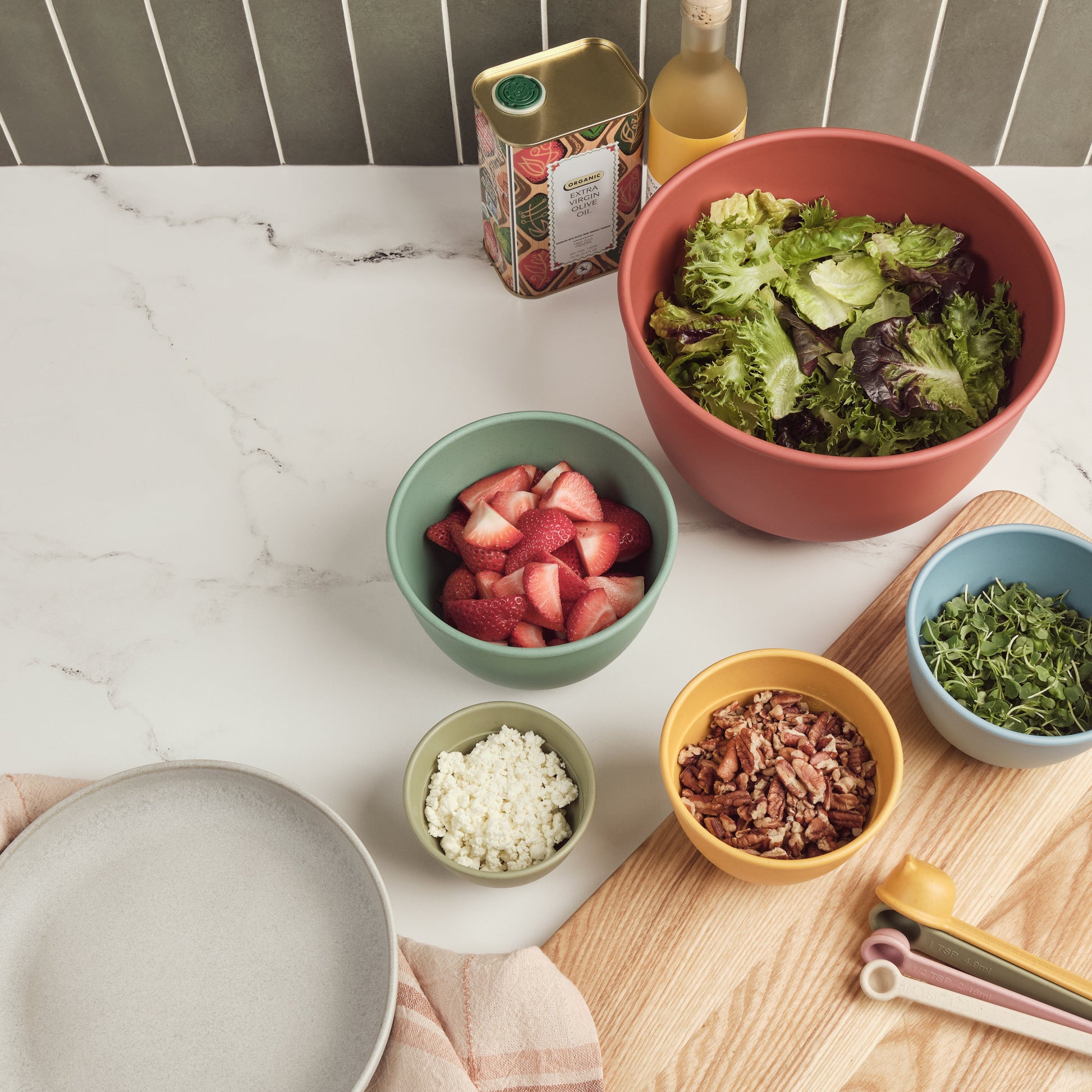 Assorted Astrik nesting bowls, measuring cups and spoons with salad ingredients on a marble countertop