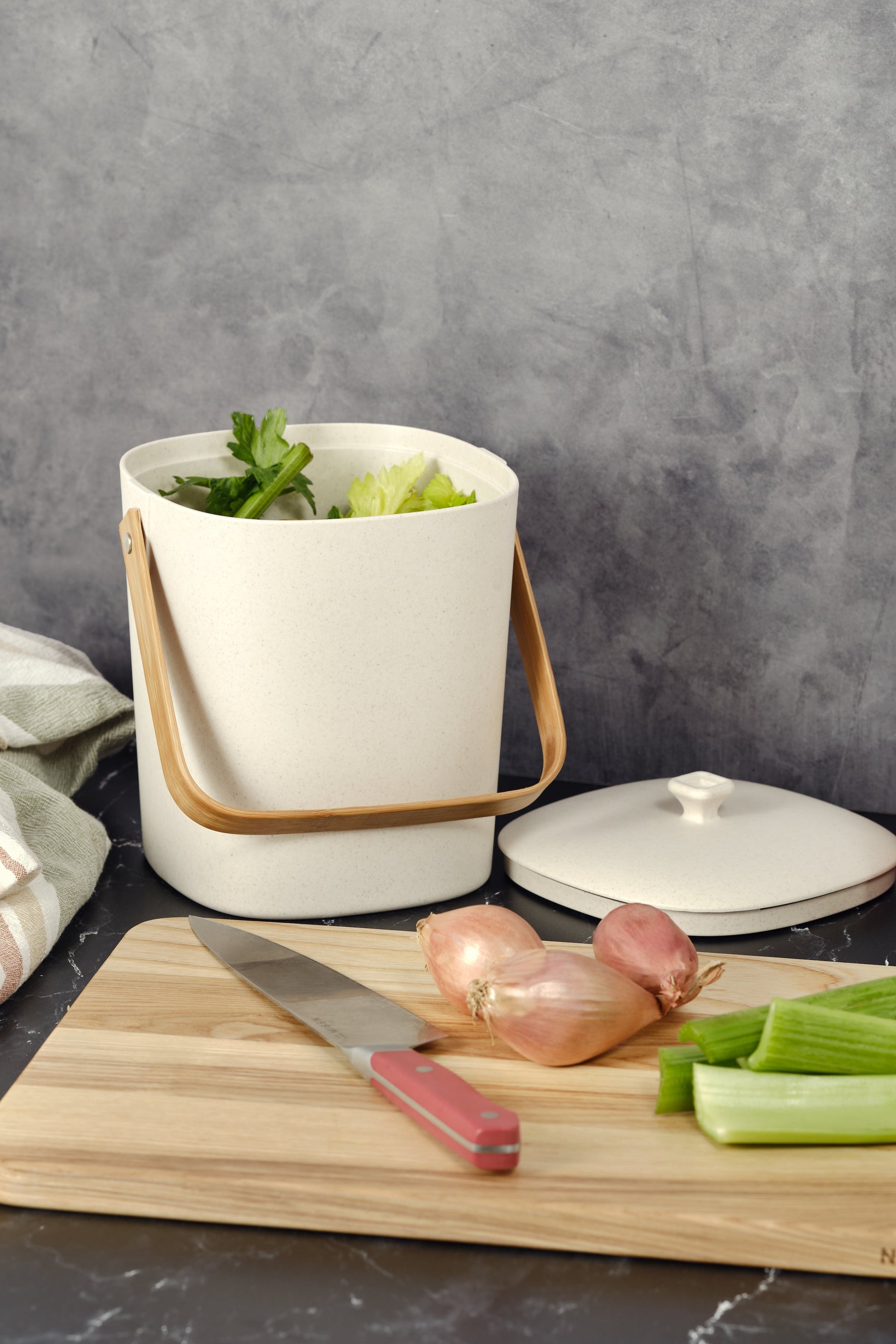 Natural-colored Astrik Compoast Bin made of 100% biodegradable plant-based plastic with a bamboo handle on a gray counter alongside a cutting board with vegetables.