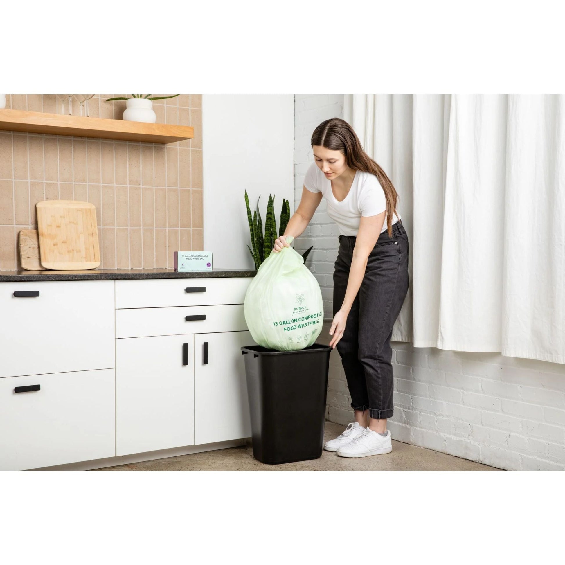 Person in a kitchen replacing a 13-gallon compostable trash bag in a black trash can.