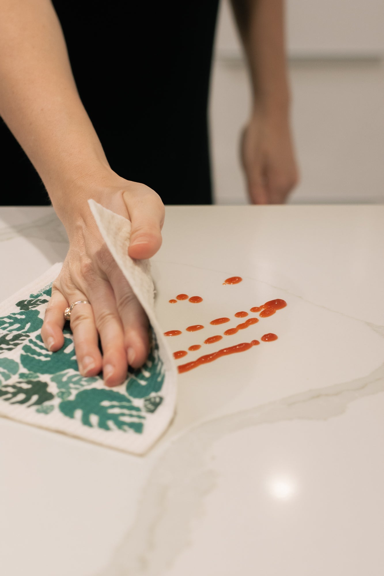Person cleaning a surface with a reusable Washcloud dish cloth, with a floral patterned towel underneath.