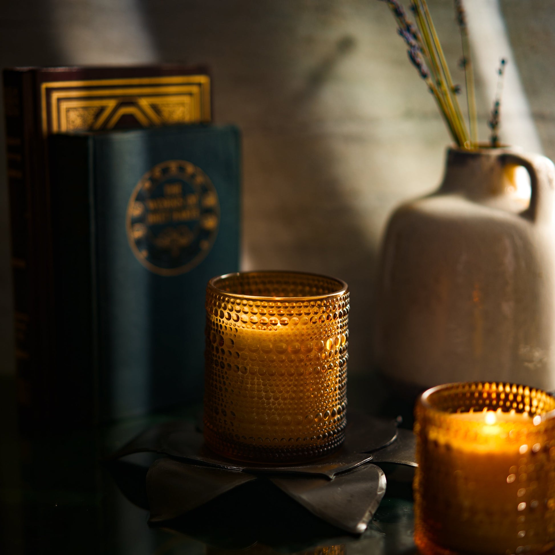Two list eco-friendly Bluecorn Aromatica Winter Scented candles in textured yellow glass holders on a dark surface with books, a white vase, and a blurred background
