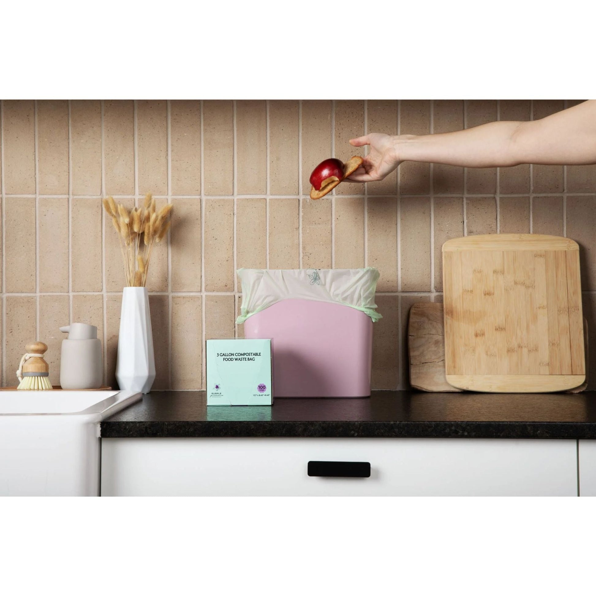 Hand dropping a red apple into a Purple Plumeria compostable kitchen garbage bag on a wooden cutting board in a kitchen.