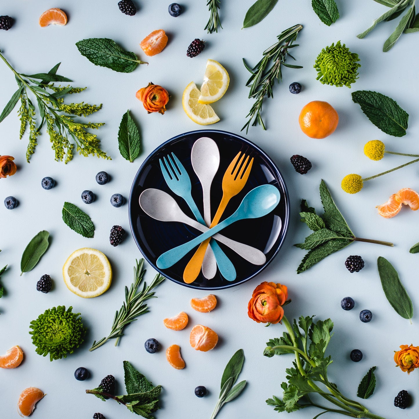 Colorful plastic-free cutlery set on a black plate surrounded by fruits, vegetables, and herbs on a light blue background. Forks and spoons are made with bamboo and compost within 2 years.