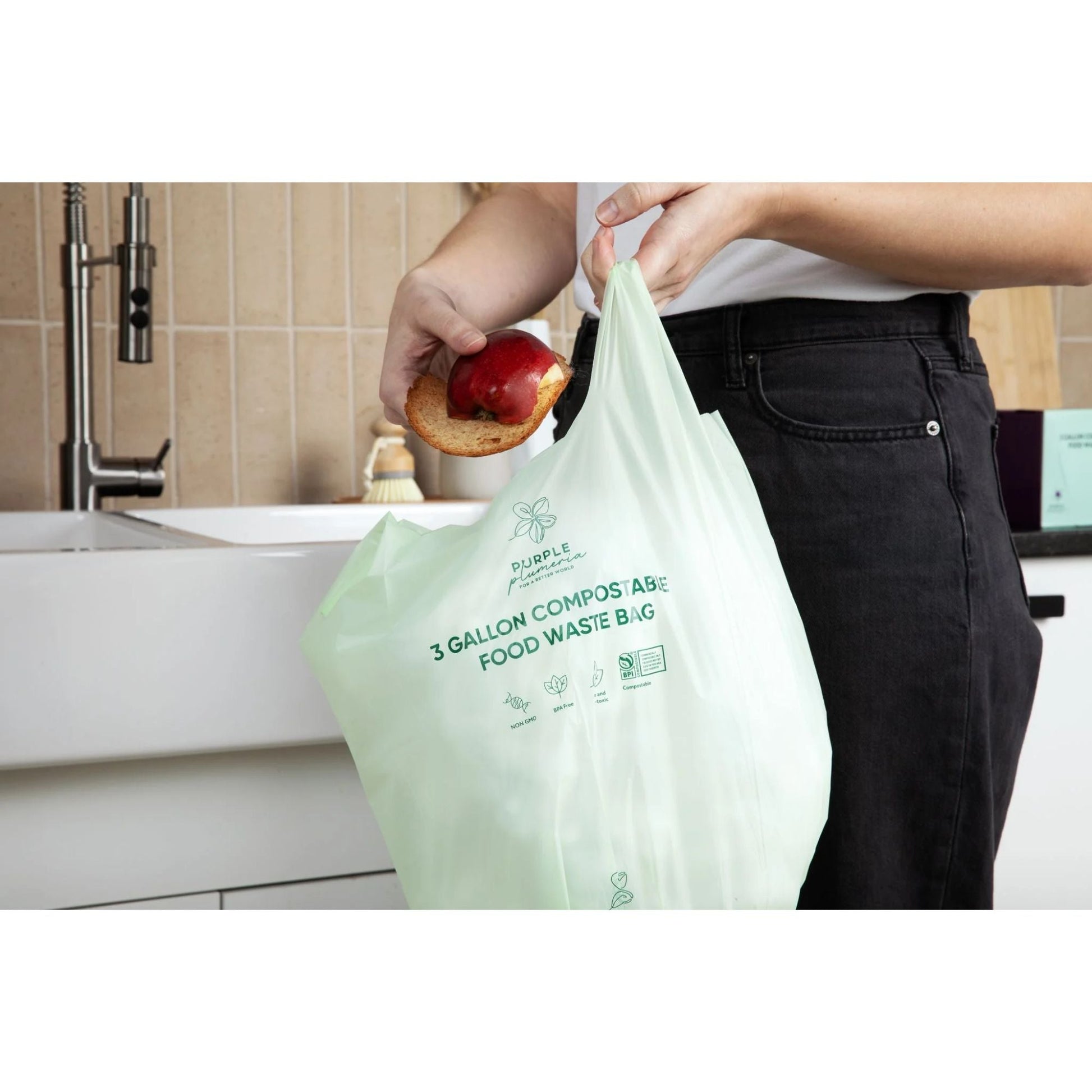 Person holding a 3 gallon Purple Plumeria compostable food waste bag with a piece of bread and apple in a kitchen.
