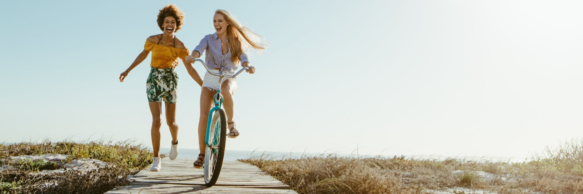 Two women, one walking, one riding a bicycle on a path near a beach.