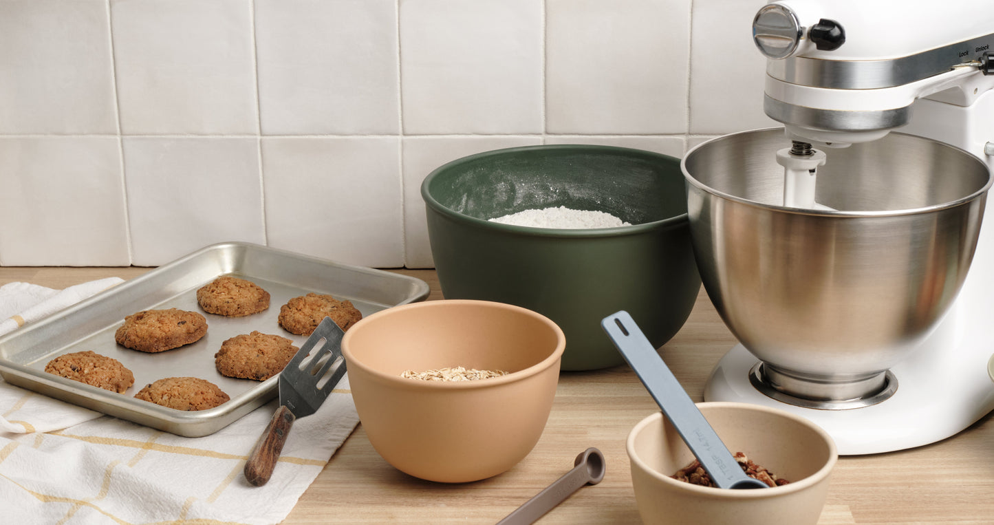 Kitchen scene with Astrik plant-based plastic nesting bowls, measuring cups and spoons with baking ingredients alongside a stand mixer and tray of cookies on a countertop.