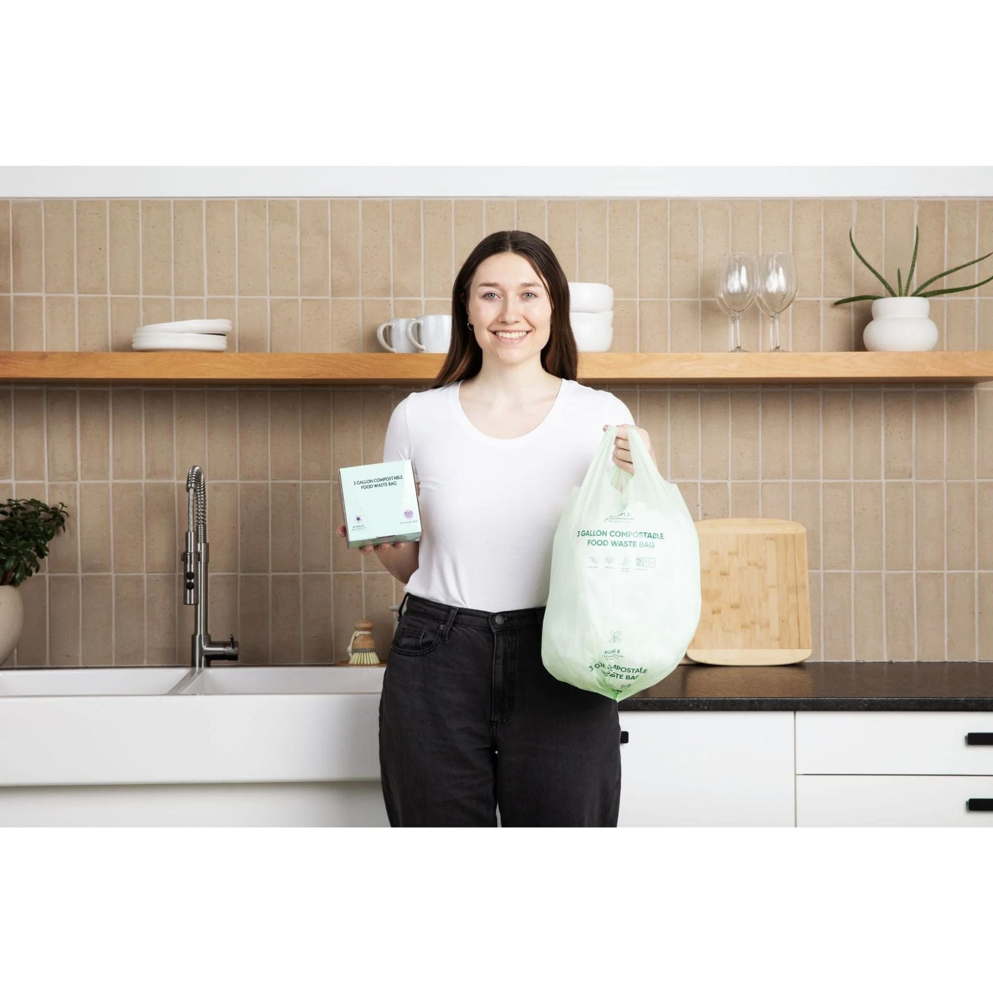 Woman holding a box of Purple Plumeria compostable garbage bags and an individual bag in a kitchen.