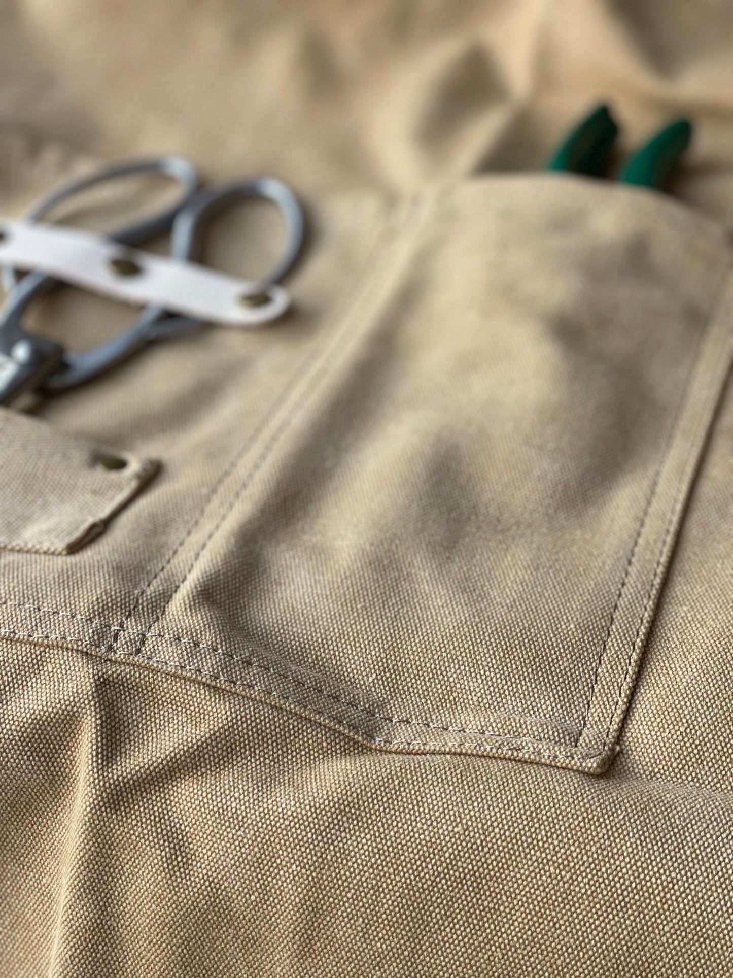 Close-up of beige garden apron made of waxed canvas with florist scissors and green garden pruner in pockets