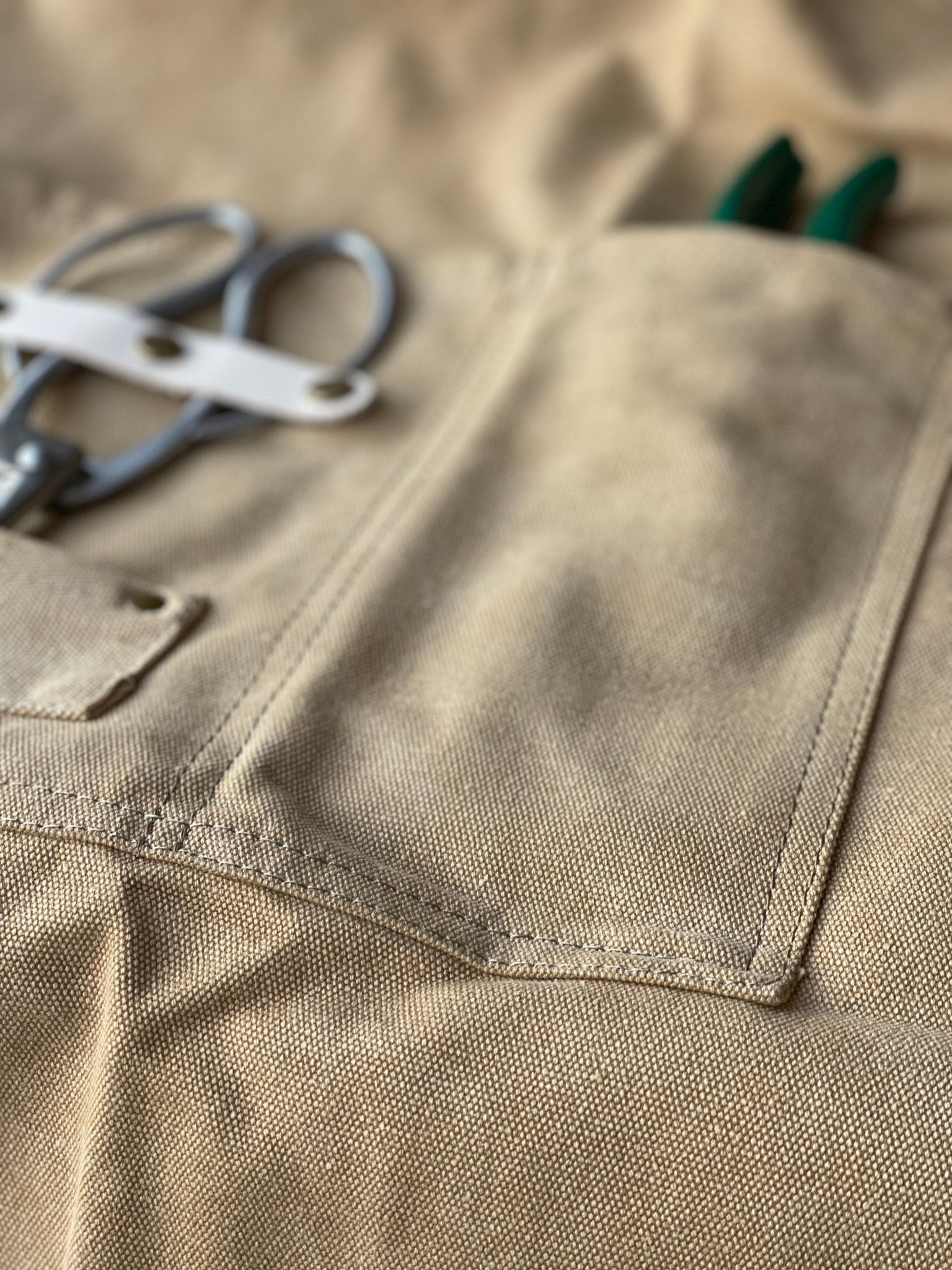 Close-up of beige garden apron made of waxed canvas with florist scissors and green garden pruner in pockets