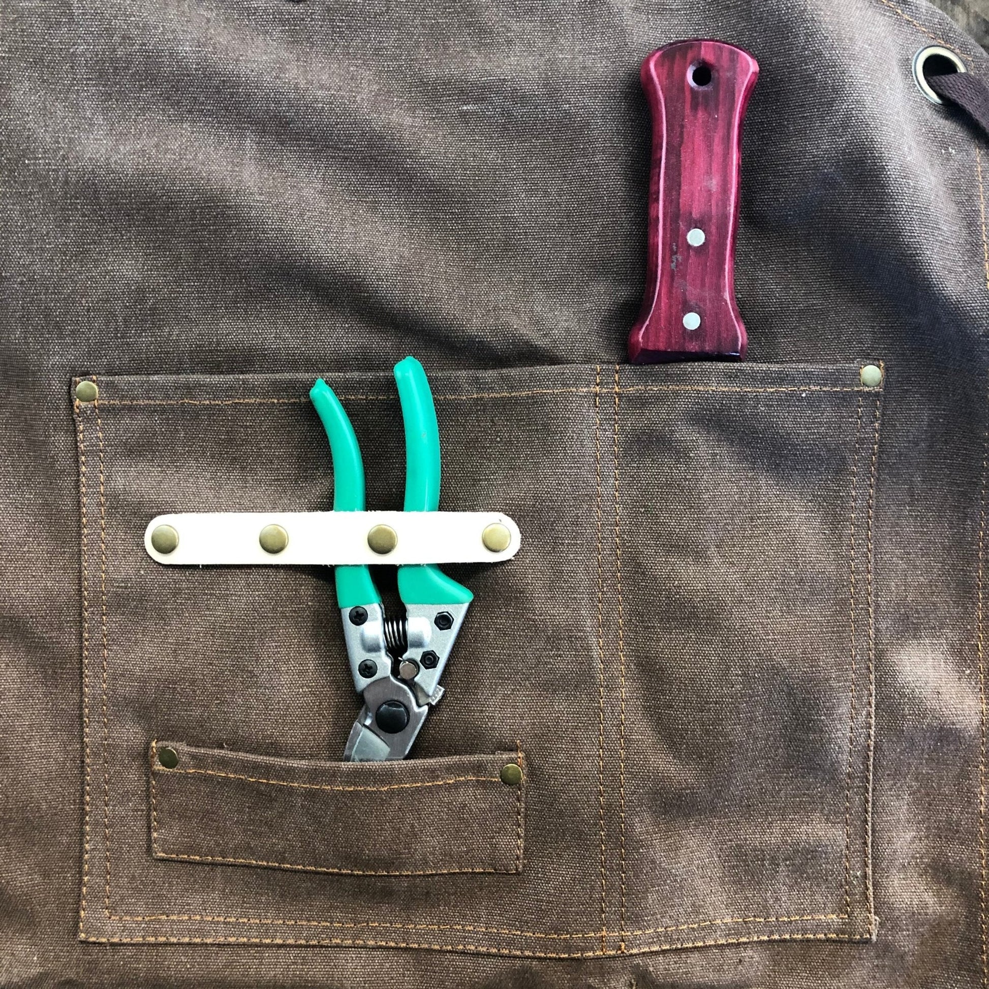 Brown leather florist apron with a pockets containing green pruning sheers and a wooden garden knife.