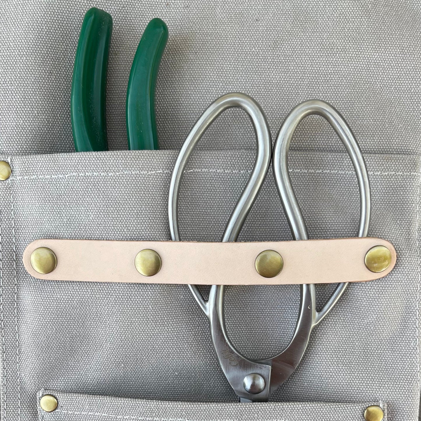 Gray tool pouch with a leather strap holding scissors and green pliers on a gray background