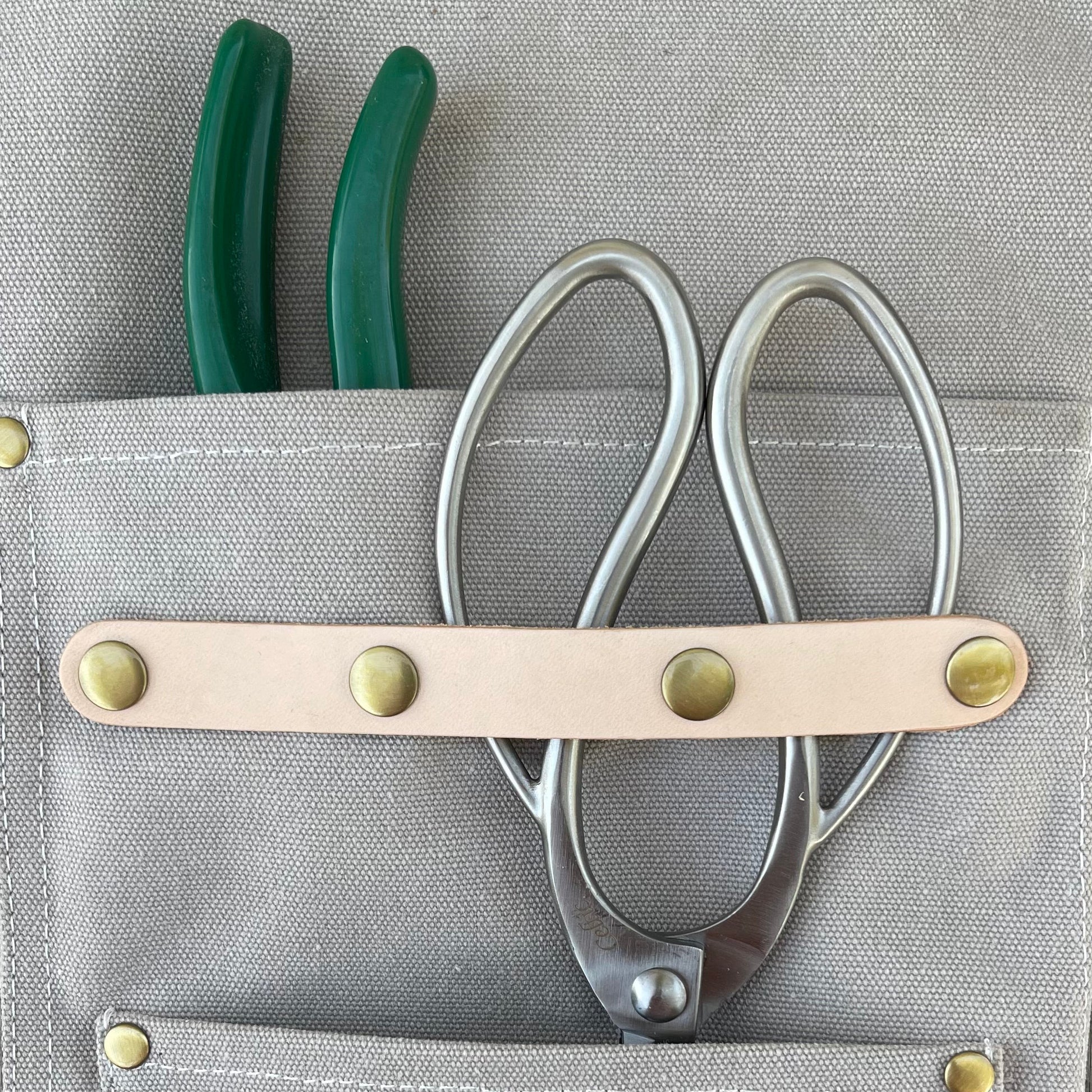 Gray tool pouch with a leather strap holding scissors and green pliers on a gray background