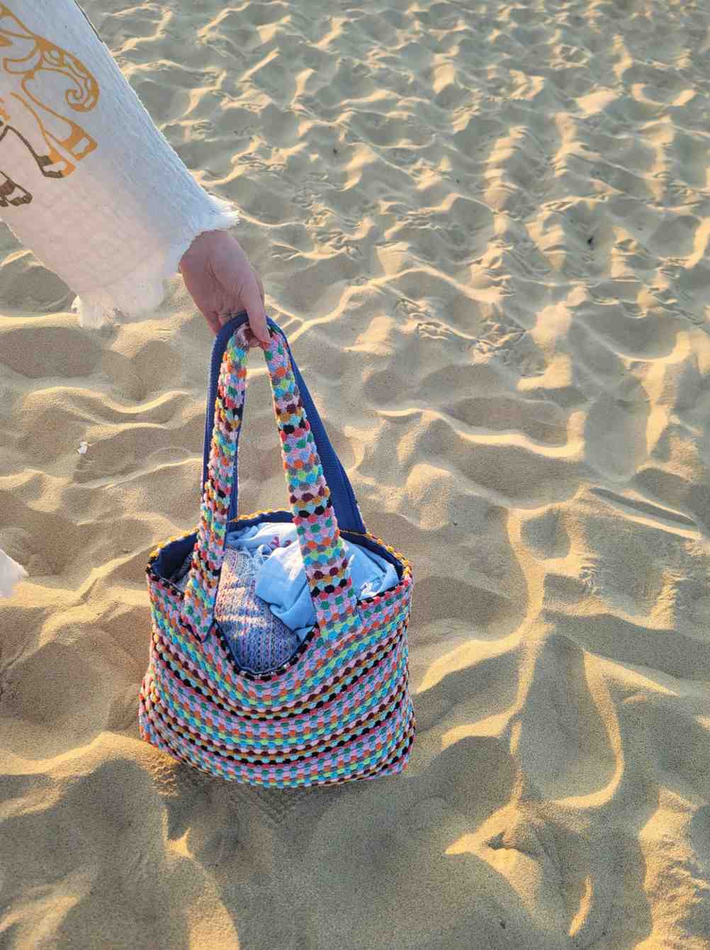 Person holding a colorful Handwoven Turkish Cotton Beach Tote – Artisan-Made & Travel Ready - on a sandy beach with water in the background