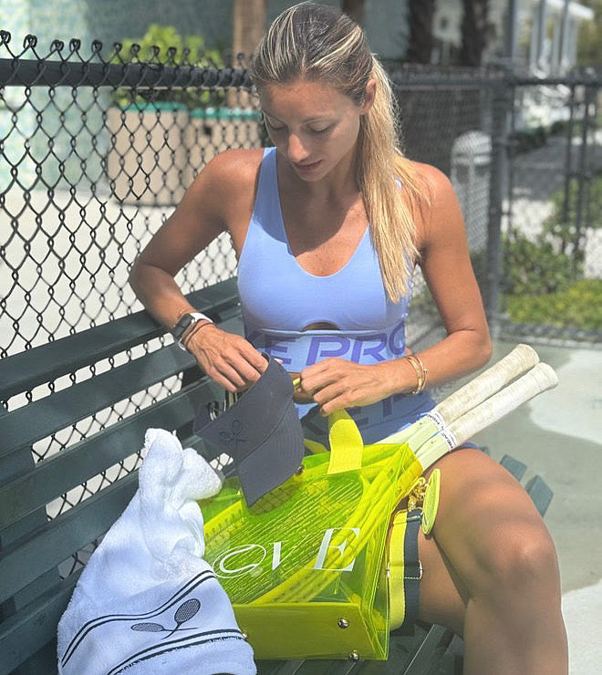 LOVE tote carrying a tennis racquet, ideal for courtside and daily use. Woman sitting on a bench with sports equipment, including a yellow bag and tennis racket.