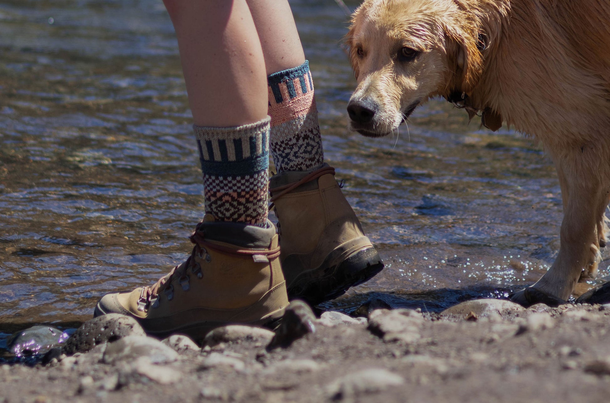 Person wearing patterned Madrona crew socks by Solmate, made with a blend of recycled merino wool, and brown boots standing near a dog by a body of water.