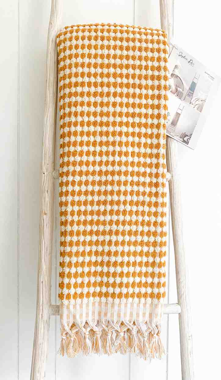 Mustard and white checkered bath towel with tassels on a wooden ladder against a white wall.