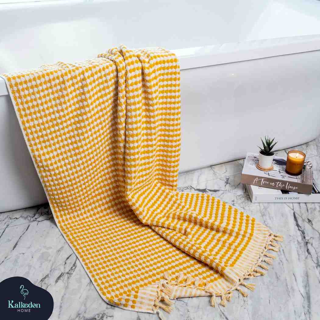 Mustard Premium Organic Turkish Jacquard Bath Towel draped over a bathtub with a marble floor and books in thejbackground.